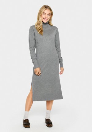 Langärmliges, graues Rollkragenkleid aus weichem Stoff, mit einem seitlichen Schlitz und gerippten Bündchen. Das Model trägt dunkle Schuhe und helle Socken.