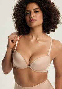 Beżowy biustonosz z wyściółką, z regulowanymi ramiączkami, bezszwowy design i gładka faktura. Modelka trzyma jedno ramiączko, prezentując kształt biustonosza.