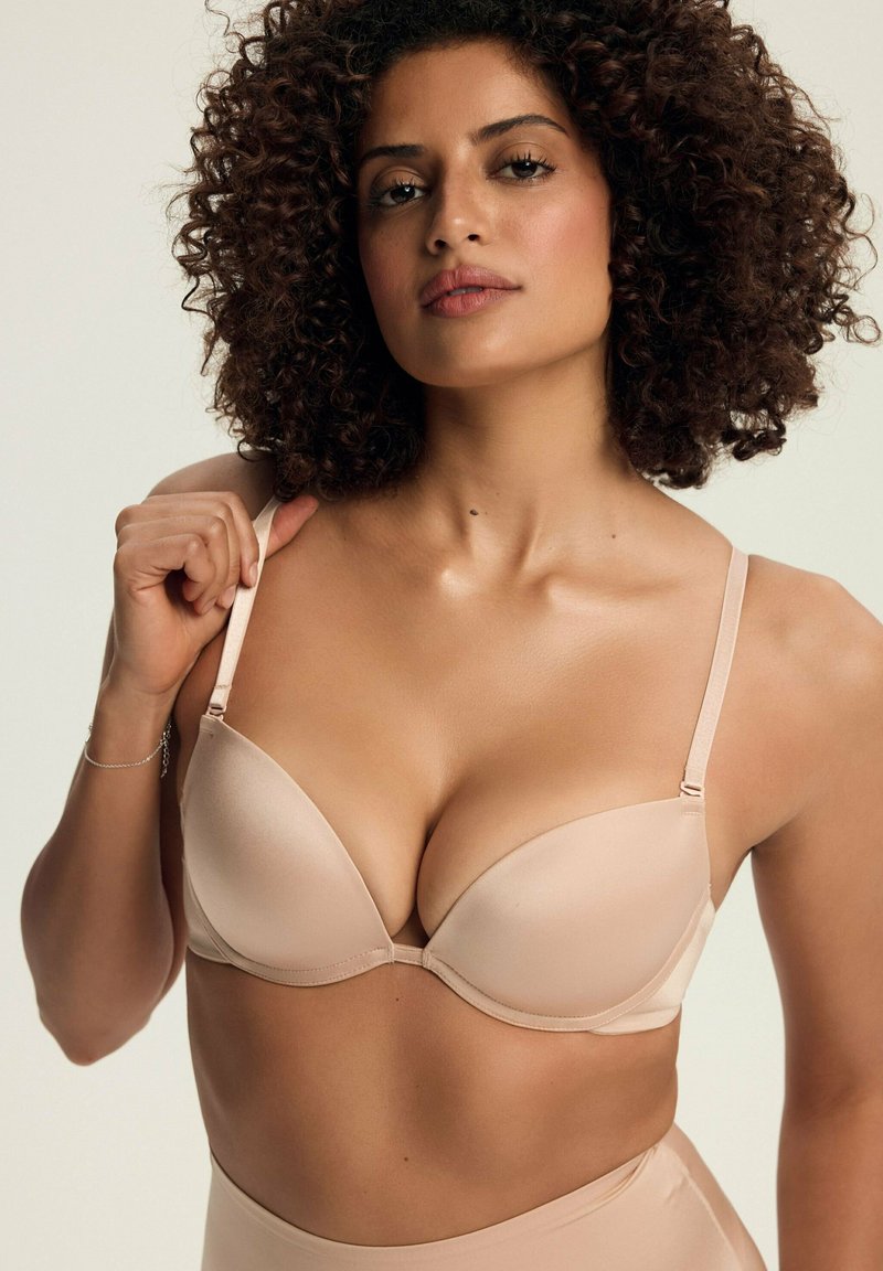 Beżowy biustonosz z wyściółką, z regulowanymi ramiączkami, bezszwowy design i gładka faktura. Modelka trzyma jedno ramiączko, prezentując kształt biustonosza.