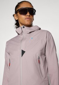 Veste imperméable rose clair avec un zip sur le devant, des accents en maille noire et deux poches latérales zippées avec des éléments en rouge.