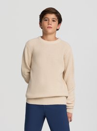 Beżowy sweter wykonany na drutach z okrągłym dekoltem, ściągaczami na mankietach i na dole. Nosi się go na niebieskie spodnie, cechuje się fakturowaną tkaniną i luźnym krojem.