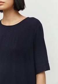 Kvinna med kort svart hår som bär en enkel mörk marinblå kortärmad topp framför en ljusgrå bakgrund.