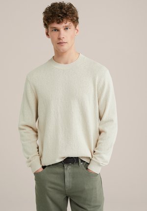 Pull beige clair avec un motif texturé, col rond et manches longues, associé à un pantalon vert olive, les mains dans les poches.