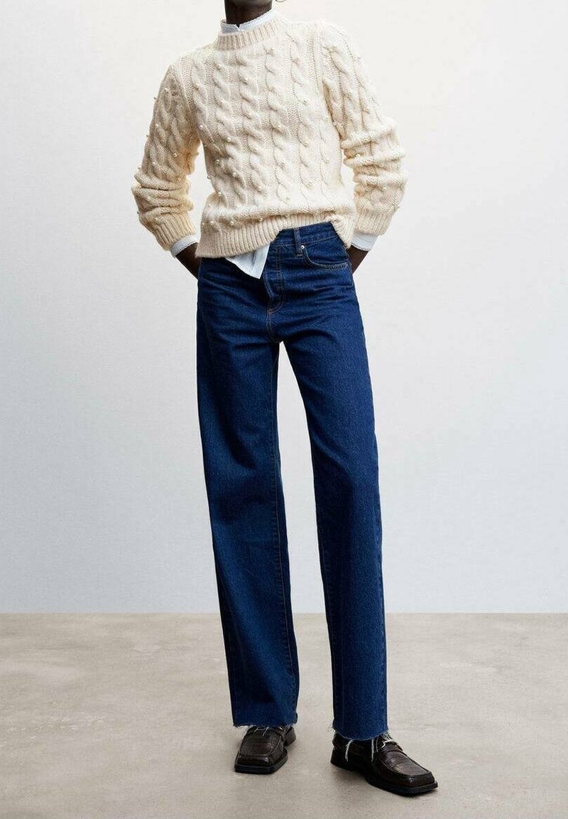 Pull en maille torsadée crème associé à un jean bleu taille haute à jambes droites et des mocassins marron foncé. Tissu texturé et coupe décontractée.