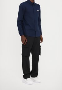 Camicia blu navy con colletto classico, abbinata a pantaloni cargo neri con tasche laterali e sneakers bianche.