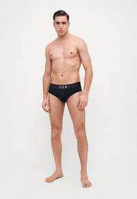 Schwarze Unterhosen mit einem elastischen Bund und einem grauen Logo. Glattes Material, taillierte Passform und niedriger Schnitt. Für Komfort geformt.