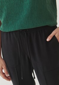 Grüner strukturierter Oberteil kombiniert mit schwarzen Hose mit elastischem Bund, Kordelzug, Seitentaschen und glatter Stoffoberfläche.