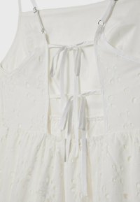 Robe blanche sans manches avec broderie florale, bretelles réglables et deux nœuds noués dans le dos ouvert avec détail taille froncée.