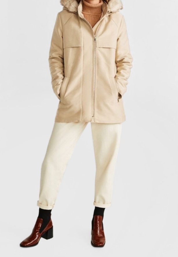 Manteau d'hiver beige avec capuche doublée en fausse fourrure, fermeture à glissière et deux poches avant, associé à un pantalon crème et des bottes à talons marron brillant.