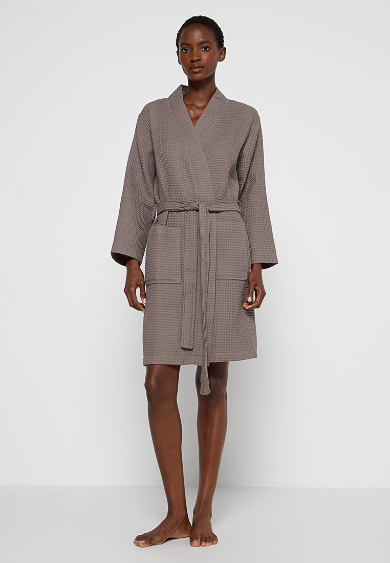 Robe marron à texture gaufrée, longueur genou, avec un design enroulé, des manches longues, une ceinture à nouer et deux poches avant. Finition en tissu lisse.