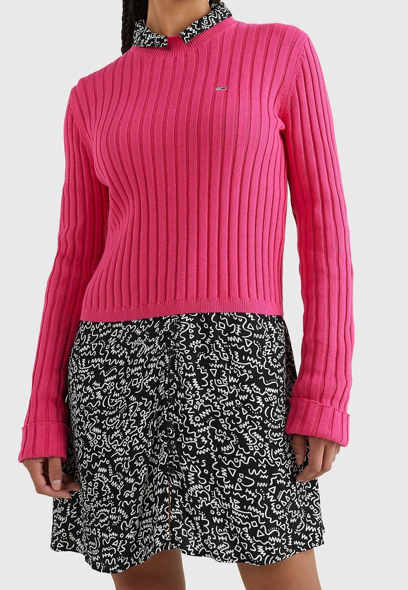 Femme portant un pull côtelé rose vif sur une jupe à motif abstrait noir et blanc avec un col de chemise assorti.
