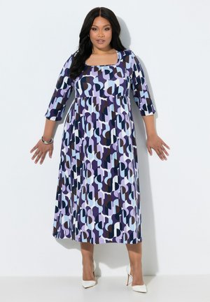 Donna che indossa un vestito midi con motivo geometrico viola, blu e nero, con maniche tre quarti e tacchi alti bianchi, in piedi contro un muro bianco.