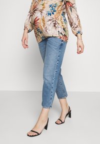 Zwangerschapsbloes met bloemenprint en lange mouwen, beige achtergrond, levendige groene, rode en gele patronen, gecombineerd met lichtblauwe cropped jeans en zwarte hakken.