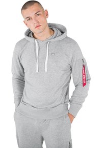 Sudadera gris hecha de tejido suave, con un bolsillo canguro en la parte frontal, cordones blancos y una etiqueta roja en la manga con texto.