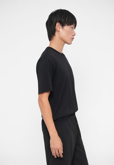 Melnas īsām piedurknēm T-krekls, izgatavots no mīksta auduma, ar apaļu kakla izgriezumu un brīvu piegriezumu, valkāts kopā ar atbilstošām melnām biksēm.