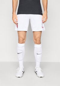 Shorts de soccer blancs avec des accents rouges et un logo ; t-shirt de sport noir ; chaussettes blanches avec le logo Nike noir ; chaussures blanches avec des détails noirs.