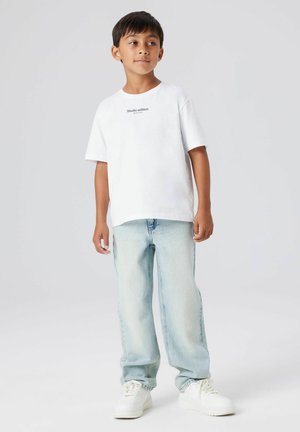 Witte katoenen T-shirt met de tekst "Studio edition", gecombineerd met lichtblauwe spijkerbroek en witte sneakers. Model staand tegen een effen achtergrond.