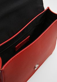 Červená kožená kabelka s hladkým povrchem, která má magnetické zapínání a černou textilní podšívku včetně kapsy na zip.