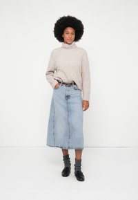 Kvinde med afrofrisure iført en beigefarvet turtleneck sweater, lys blå denim midi-nederdel, grå sokker og sorte loafers, der står foran en hvid baggrund.