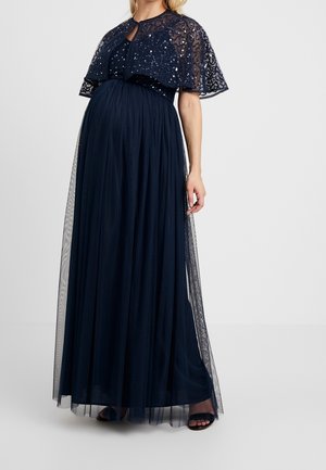 Gravid kvinde iført en gulvlængde mørkeblå kjole med en palietbesat kappe og gennemsigtigt lag, stående med den ene hånd afslappet ved siden.