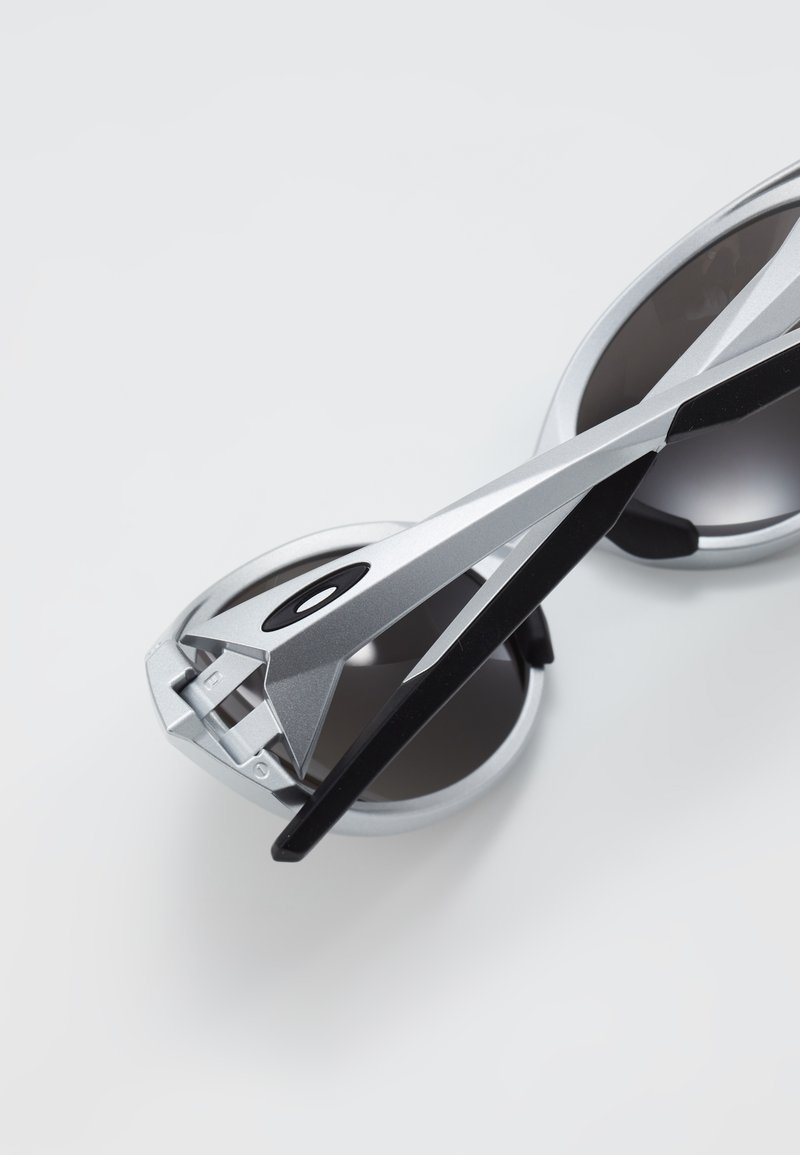 Zonnebril met een zilveren frame, zwarte accenten en donkere glazen. Hoekig ontwerp met kenmerkende pootjes, die een robuuste afwerking van het materiaal laten zien.