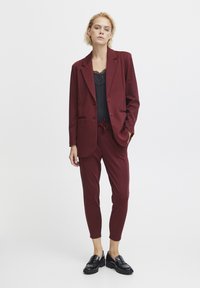Burgundský blejzer s klopami a gombíkovým zapínaním, skombinovaný s nohavicami taperovaného strihu a čiernou čipkovou kamisolou. Čierne loafers dotvárajú vzhľad.