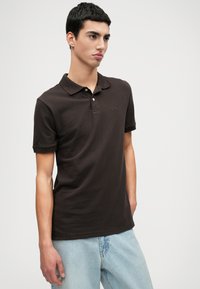 Σκούρο καφέ πόλο μπλουζάκι με κοντά μανίκια, γιακά και τρία κουμπιά μπροστά. Διακριτό λογότυπο στο στήθος. Συνδυασμένο με ανοιχτό μπλε τζιν παντελόνι.