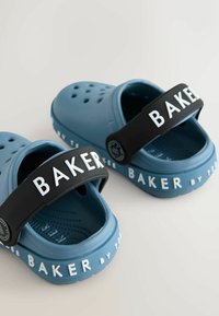 Modré gumové klobúky s ventilačnými otvormi, čierny popruh s nápisom "BAKER" v bielej farbe, textúrované vložky a branding "by T" na boku.