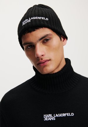 Black ribbed beanie features white embroidered "KARL LAGERFELD JEANS" logo. The model wears a matching black turtleneck sweater.