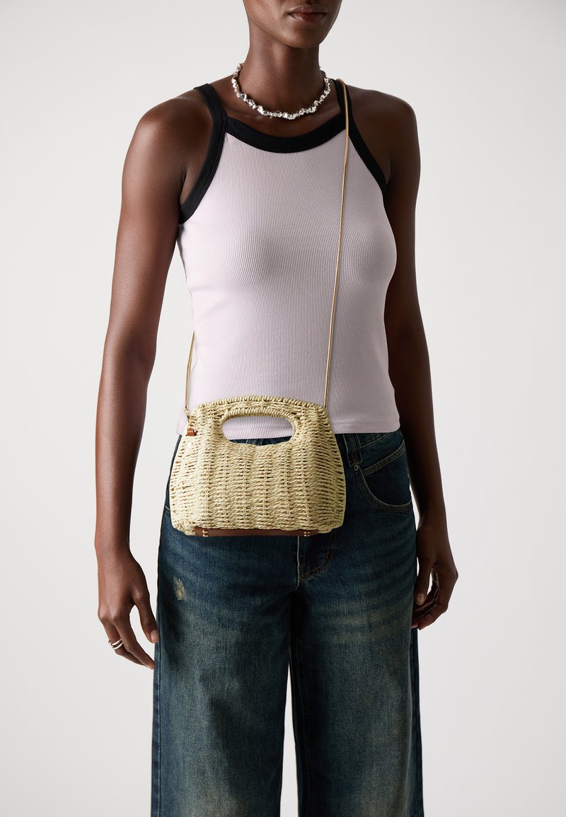 Gewebte Strohtasche mit rundem Deckel und einem einzelnen Schultergurt. Beigefarben mit einer glatten Textur und subtilen Details an den Kanten.