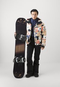 Snowboard s crnim i drvenim uzorkom, kojeg drži osoba u šarenoj cvjetnoj jakni s podstavom, crnim hlačama i rukavicama.