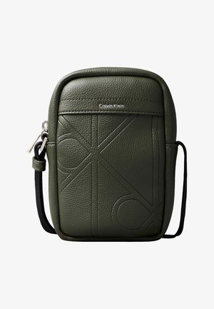 Mała oliwkowozielona torba na ramię Calvin Klein z dużymi, tłoczonymi inicjałami oraz czarnym regulowanym paskiem, zapinana na zamek po lewej stronie.