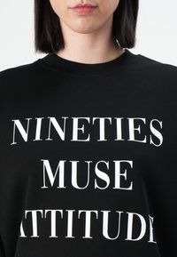 Felpa nera con testo bianco in evidenza: "ATTITUDINE MUSE DEGLI ANNI NOVANTA." Collo rotondo e tessuto morbido con un taglio rilassato.