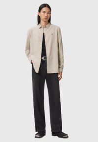 Camicia button-up beige chiaro con maniche lunghe, dettaglio del logo singolo sul petto, abbinata a jeans larghi scuri e scarpe nere.