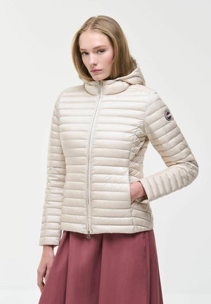 Donna con giacca trapuntata beige chiaro con cappuccio e zip frontale e gonna color granata, in piedi con una mano in tasca.