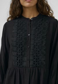 Cămașă neagră cu nasturi, având un design brodat texturat pe față, cu talie strânsă și mâneci lungi. Țesătură netedă.