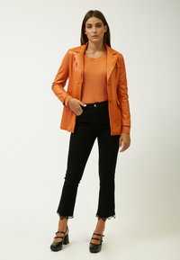 Blazer de cuero sintético naranja con solapa, top de punto naranja, vaqueros negros de campana con encaje, y zapatos de plataforma negros con correas.