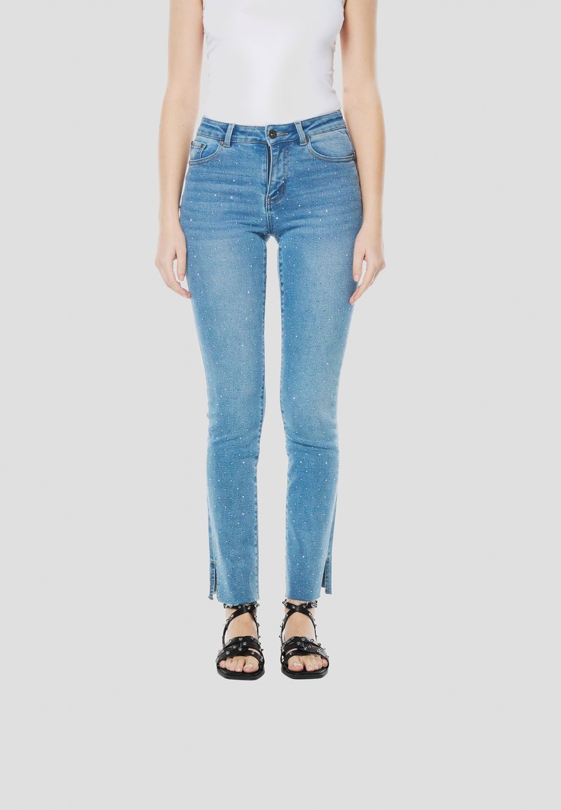 Lichtblauwe skinny jeans met decoratieve strass-steentjes, hoge taille en zijopeningen bij de zoom, gecombineerd met zwarte sandalen met bandjes.