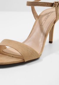 Sandale à talon haut beige en daim à bout ouvert avec bride cheville et boucle, semelle intérieure cousue, et logo Esprit sur le talon.