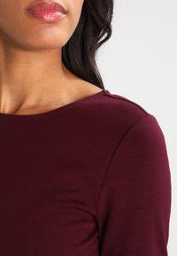 Haut bourgogne avec un col bateau classique, texture lisse et tissu doux, présentant des détails de couture subtils sur les épaules.