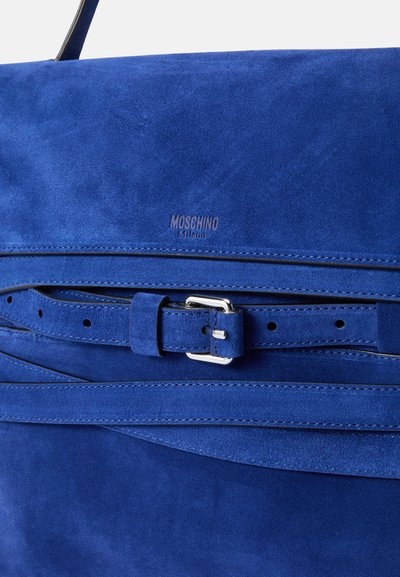 Modra suedna torbica z gladko teksturo, ki ima podrobnosti z zaponko in trakom. Vključuje embosirano oznako "Moschino Milano."
