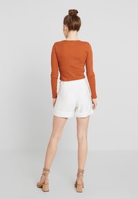 Ribbade rostfärgade långärmade toppar ihop med vita high-waisted shorts med veckad detaljering och bandade beige klackar. Enfärgad bakgrund.