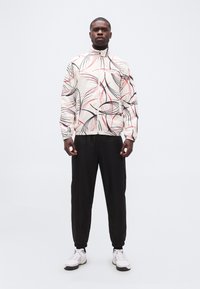 Άνδρας που φοράει λευκό μπουφάν με φερμουάρ, διακοσμημένο με μαύρες και κόκκινες αφηρημένες γραμμές, μαύρο παντελόνι και λευκά αθλητικά παπούτσια, στέκεται μπροστά σε απλό φόντο.