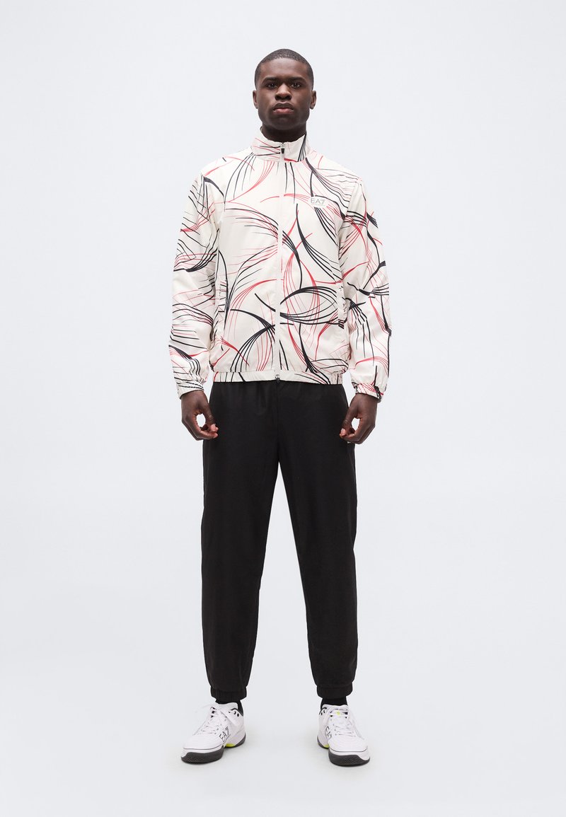 Άνδρας που φοράει λευκό μπουφάν με φερμουάρ, διακοσμημένο με μαύρες και κόκκινες αφηρημένες γραμμές, μαύρο παντελόνι και λευκά αθλητικά παπούτσια, στέκεται μπροστά σε απλό φόντο.