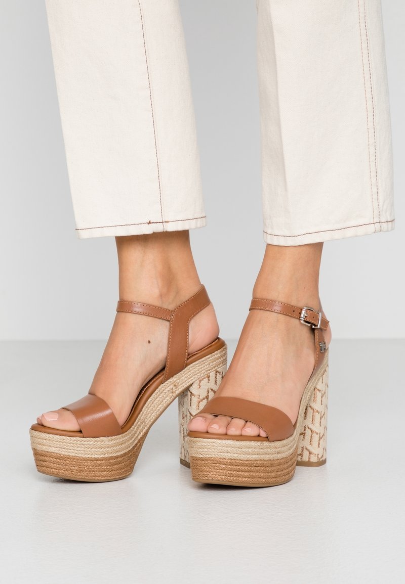 Sandales compensées en cuir fauve avec brides à la cheville et détails en corde tressée, portées avec un pantalon blanc raccourci.
