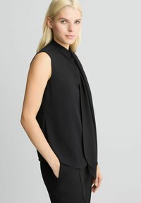 Zwarte mouwloze blouse met een hoge hals en een gedrapeerde voorkant. Soepele stof met een losse pasvorm, gecombineerd met zwarte broek.