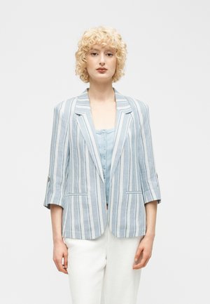 Femme aux cheveux blonds bouclés portant un blazer à rayures bleues et blanches sur un haut bleu clair boutonné et un pantalon blanc, debout devant un fond uni.
