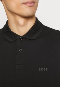 Svart polo-skjorta med en texturerad krage, treknappsslån och diskret "BOSS"-logotyp i kontrasterande färg på vänster sida.