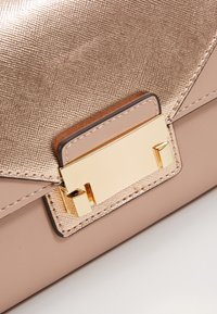 Gros plan d’un sac à main beige avec rabat texturé et fermoir rectangulaire doré brillant sur une base en cuir lisse.