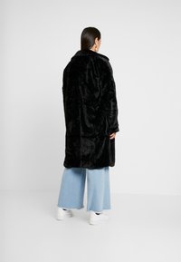 Musta tekoturkiskotelo, jossa on kaulus, sivutaskut ja rento leikkaus, yhdistettynä vaaleansinisiin leveisiin farkkuihin ja valkoisiin lenkkareihin.